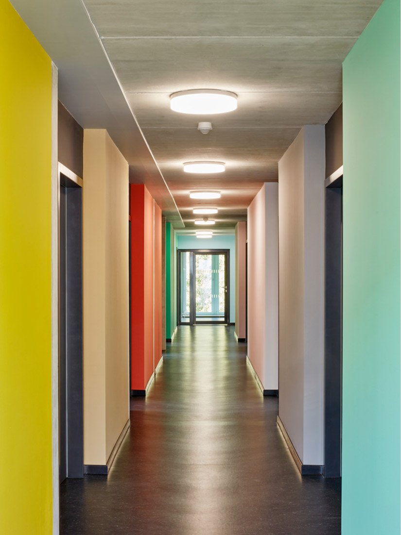 The corridors are flanked with colored wall sections and either draw the eye into the green outdoors or seem to step a little to the side to allow entry into the living spaces.
Photo © Sigurd Steinprinz / ACMS Architekten GmbH